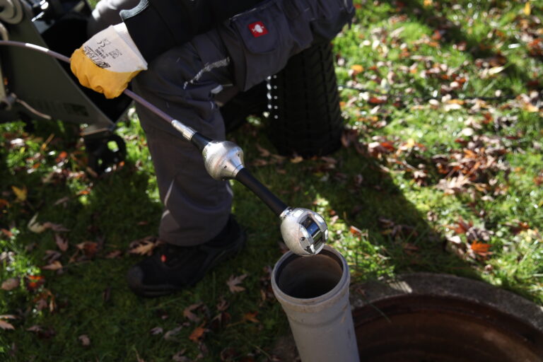 Worker in gloves and gray coveralls inserts a tool into an open PVC pipe outdoors.