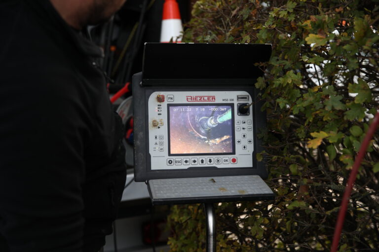Technician operating a handheld inspection controller on a tripod outdoors, with a green shrub nearby and an orange cone in view behind the device.